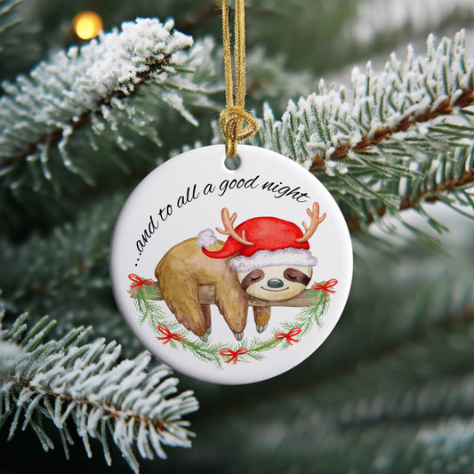 Christmas ornament that has a sleeping sloth laying on a branch wearing a Santa hat with reindeer antlers. Bottom of ornament has a Christmas garland and top of ornament has the words And To All A Good Night.